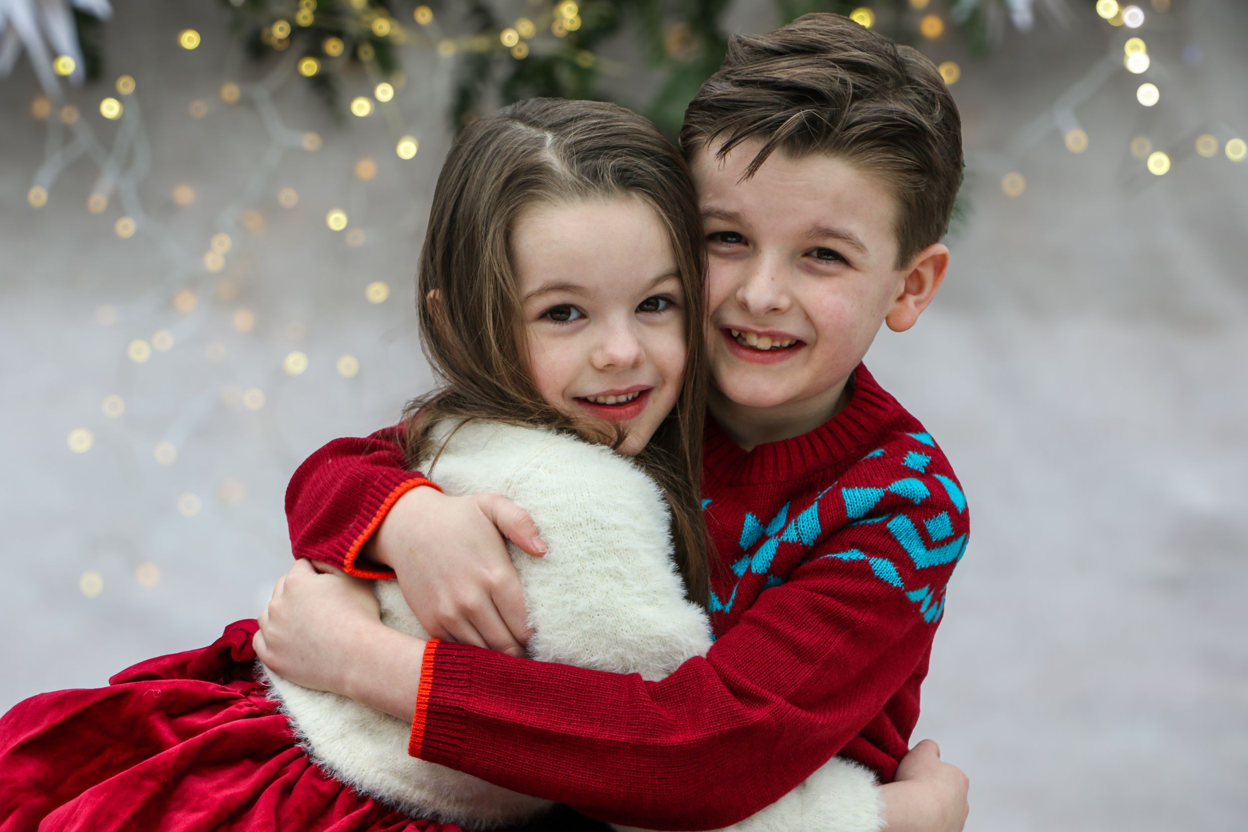 Two children in red and blue holiday sweaters embrace while smiling against a festive winter backdrop.