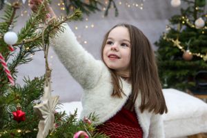 Child in white sweater and red dress decorating Christmas tree with ornaments.