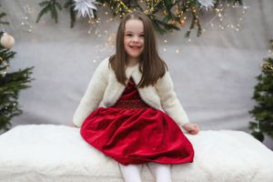 Child in red dress and white cardigan sitting on white bench with holiday decorations.
