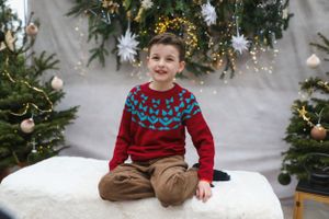 Child wearing festive red and blue patterned sweater sitting on white bench with holiday backdrop.