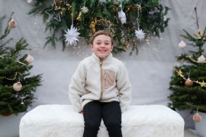 Child in cream colored fleece jacket sitting on white bench between Christmas trees.
