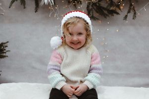 Child wearing knit hat and pastel sweater sitting on white bench with holiday decorations.