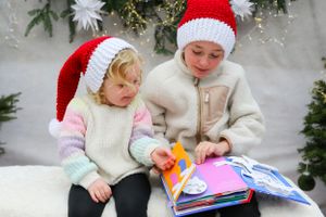 Two children in winter sweaters and Santa hats reading together on white bench.