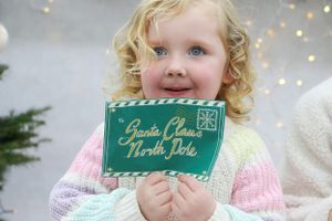 Child in pastel sweater holding green Santa Claus thank you card with sparkly backdrop.