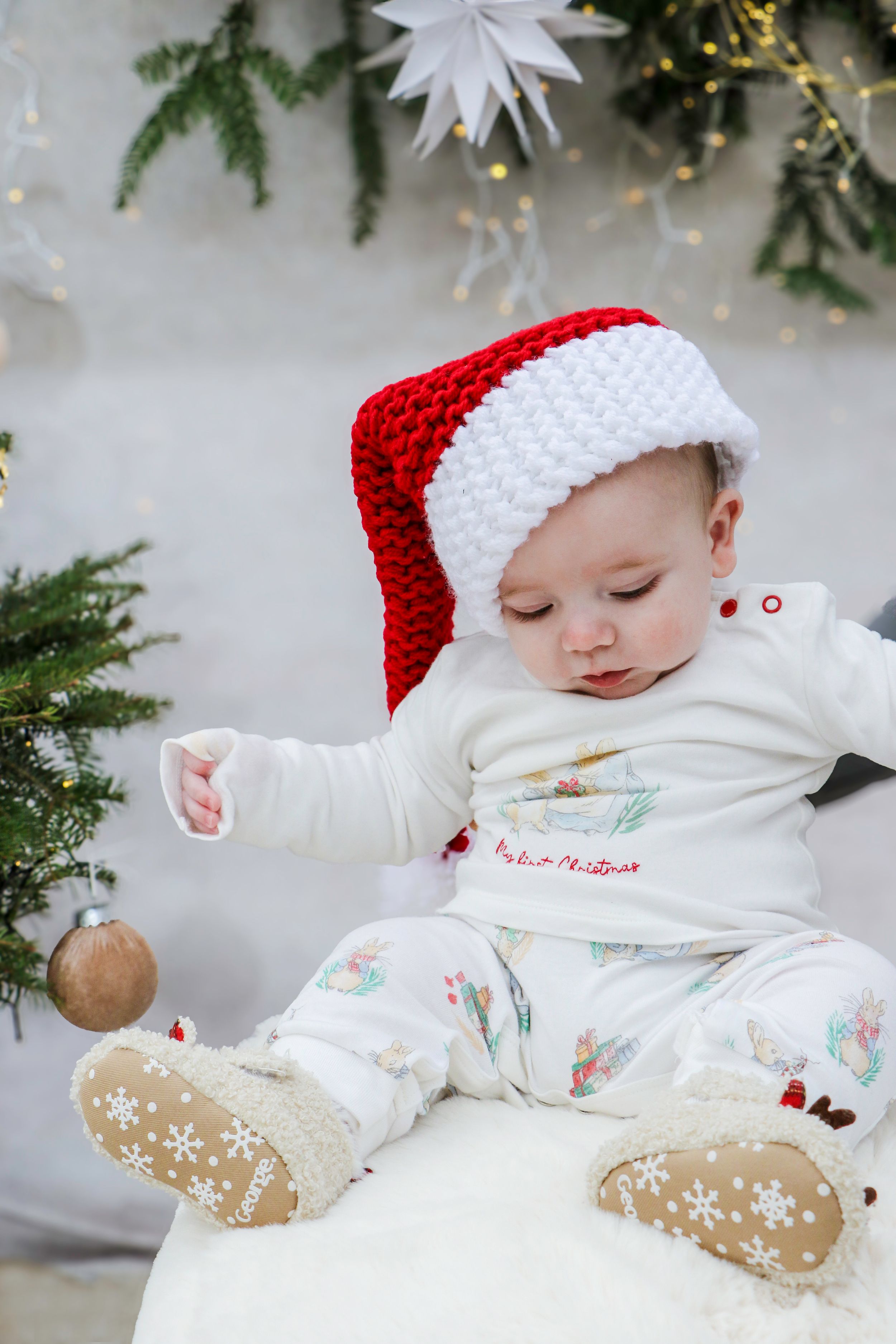 Baby wearing white outfit and red Santa hat sitting against Christmas backdrop.