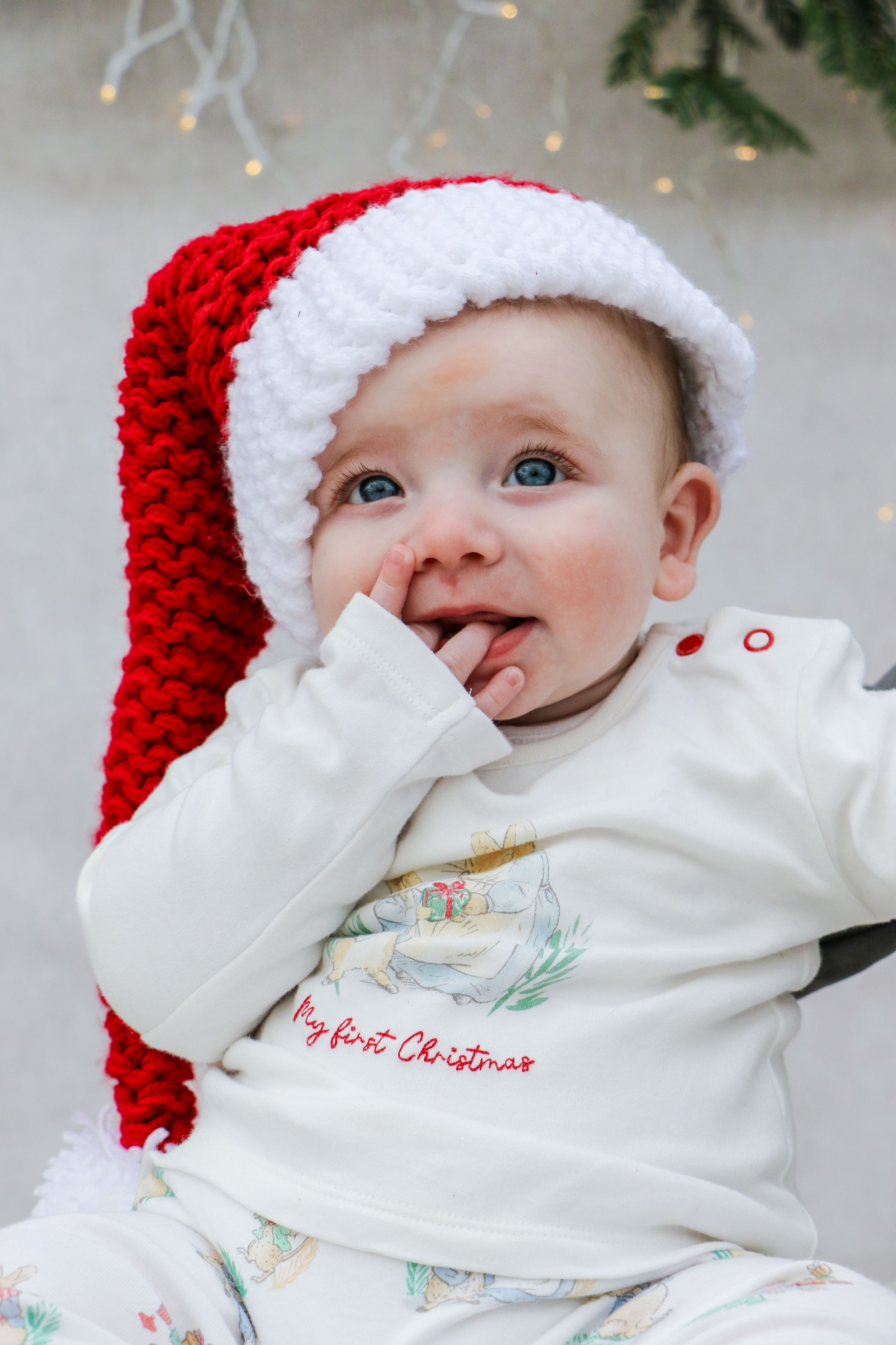 Baby in festive red and white hat with Christmas themed outfit against holiday decor.