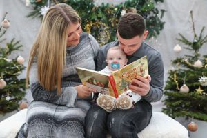 Couple sharing a Christmas storybook while cuddled under a gray blanket by decorated trees.
