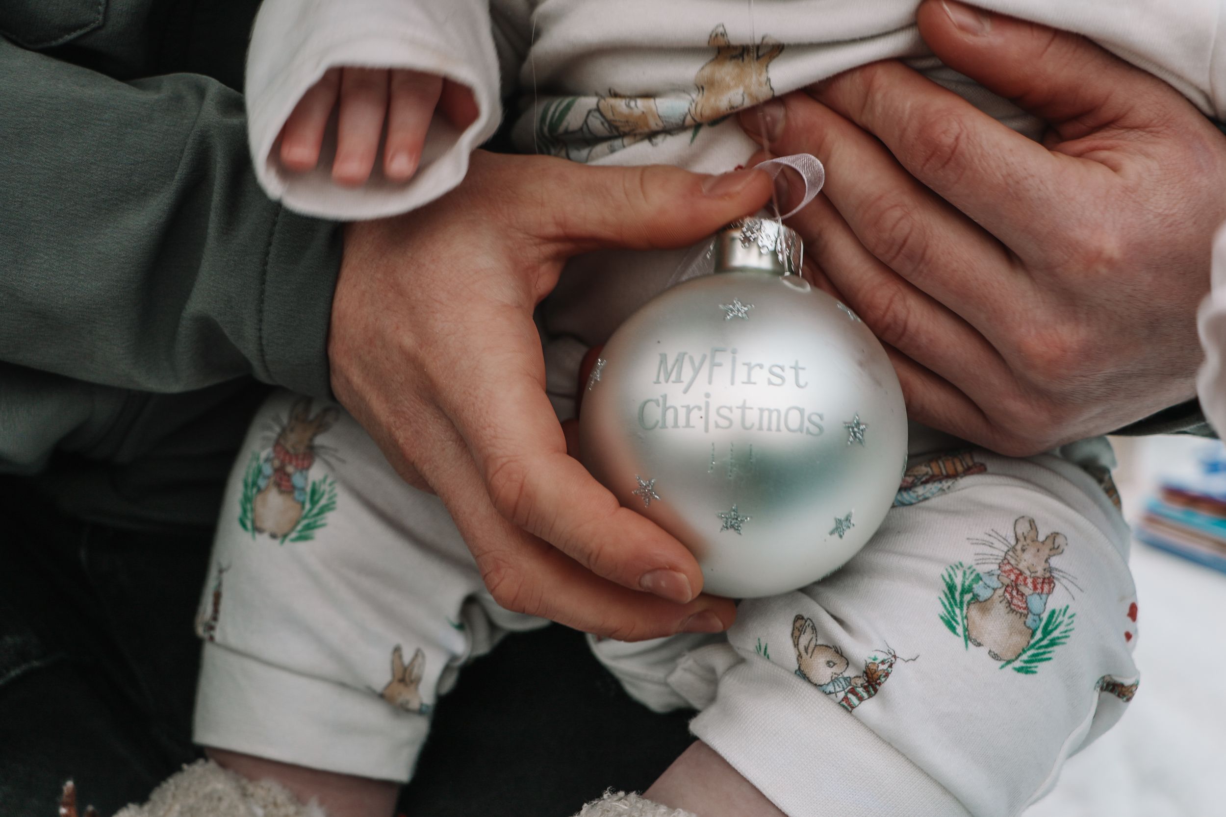 Hands holding a silver Christmas ornament ball with festive baby clothing visible.