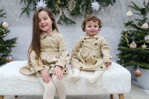 Two children in cream-colored outfits sitting on a white bench with Christmas decorations in background.