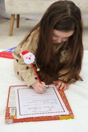 Writing a Christmas letter with Santa pen while lying on white floor near holiday decorations.