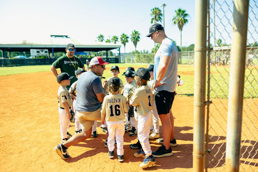 Baseball players sit on dugout bench during sunny game at outdoor sports field with palm trees in background.