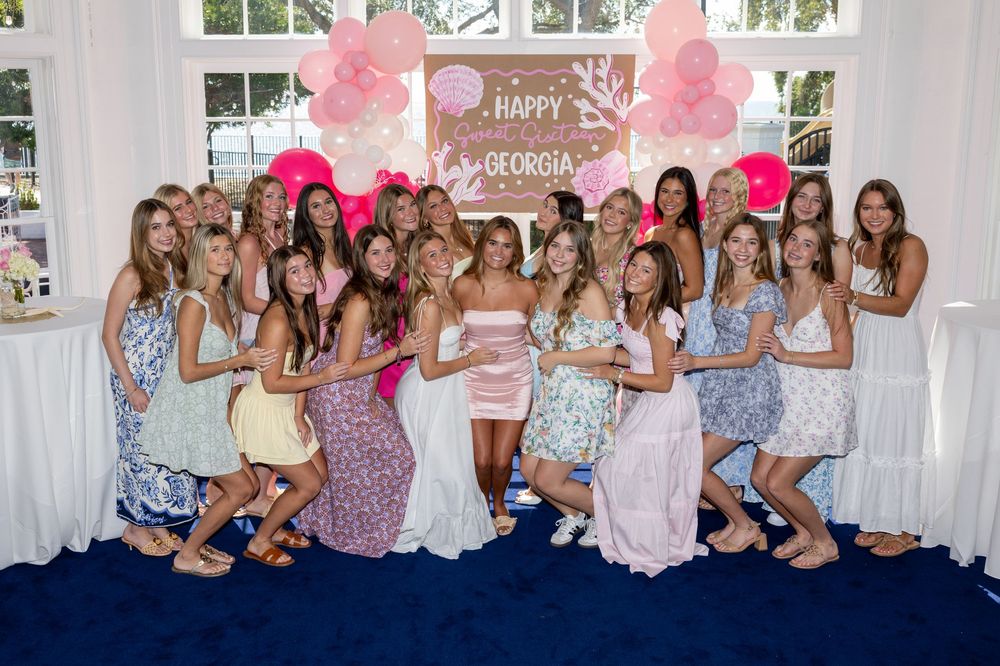A group of people pose together at a party celebration with pink balloons and floral decorations in the background.