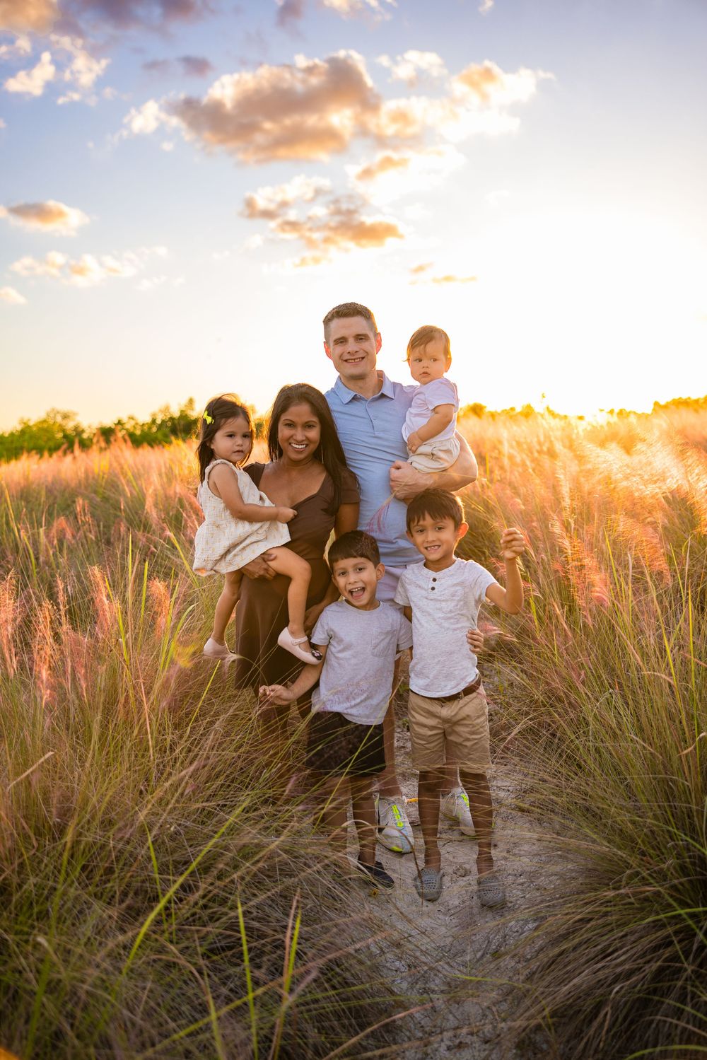 A family poses together in tall grass during golden hour sunset with warm glowing sky in the background.