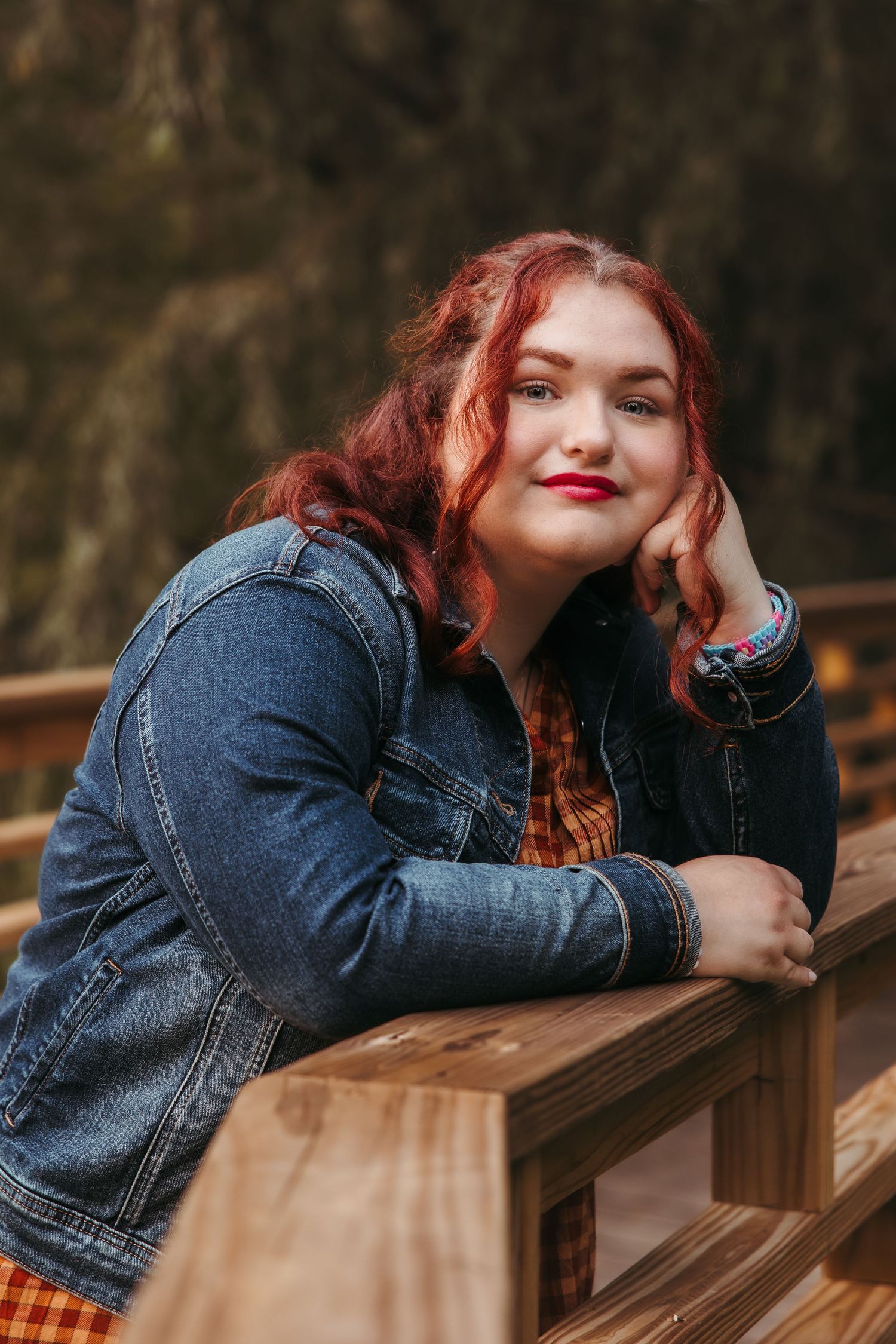 Portrait of a high school senior with vibrant red hair and bright lipstick wearing denim jacket leaning on wooden railing outdoors.