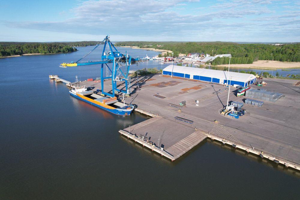 Large blue crane at a waterfront shipping terminal with cargo handling equipment.