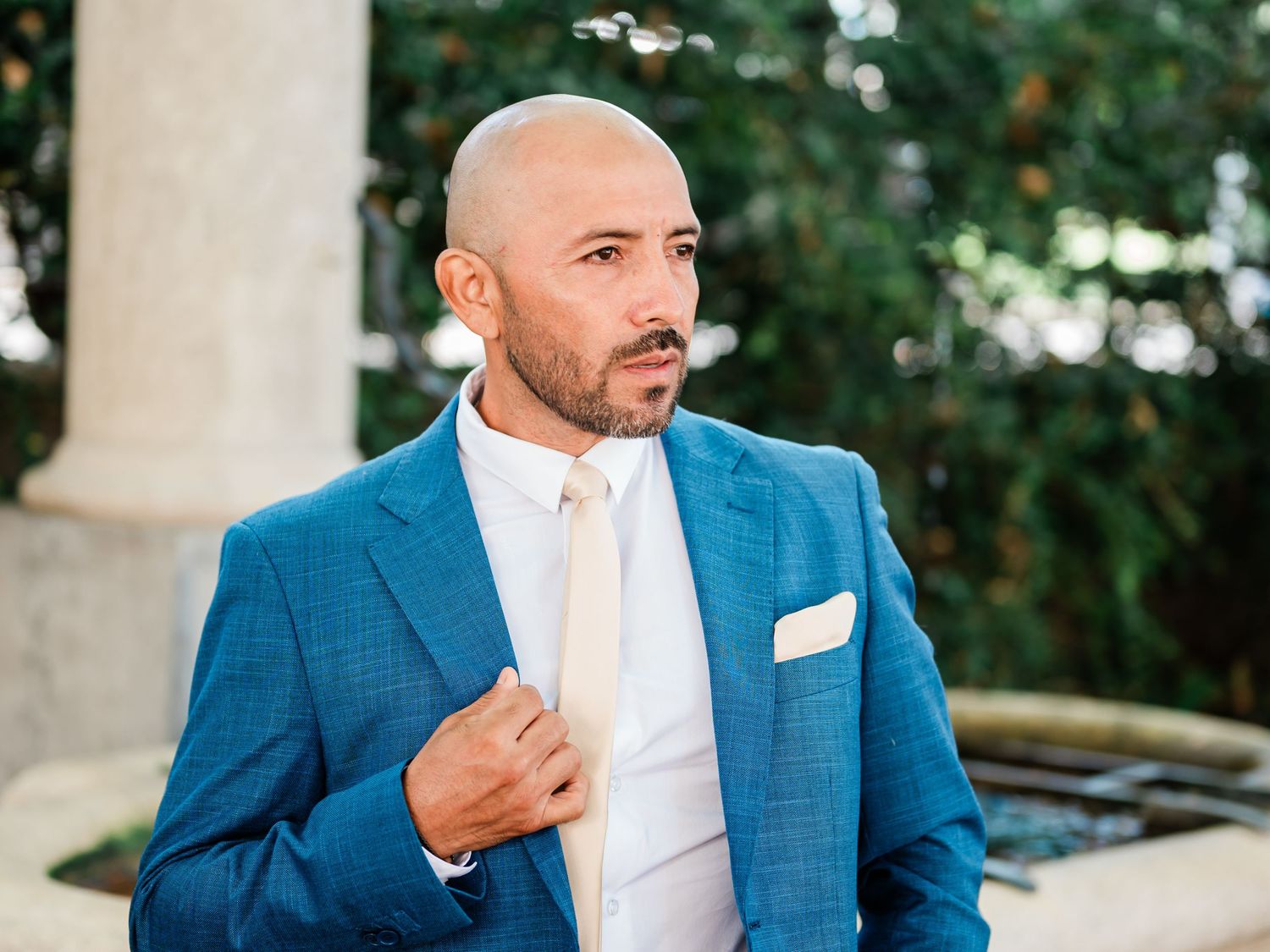 A groom in a sharp blue suit adjusts his jacket, exuding confidence and style, with a serene outdoor background framing the moment in Geneva