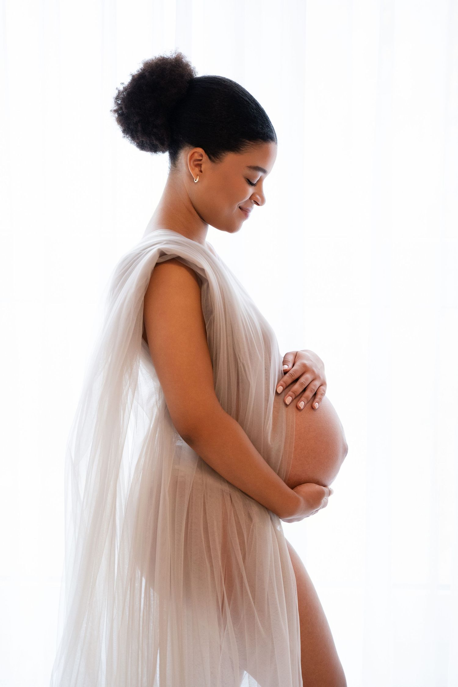 Pregnant woman in sheer beige wrap gently cradling her baby bump, smiling softly against a white background.