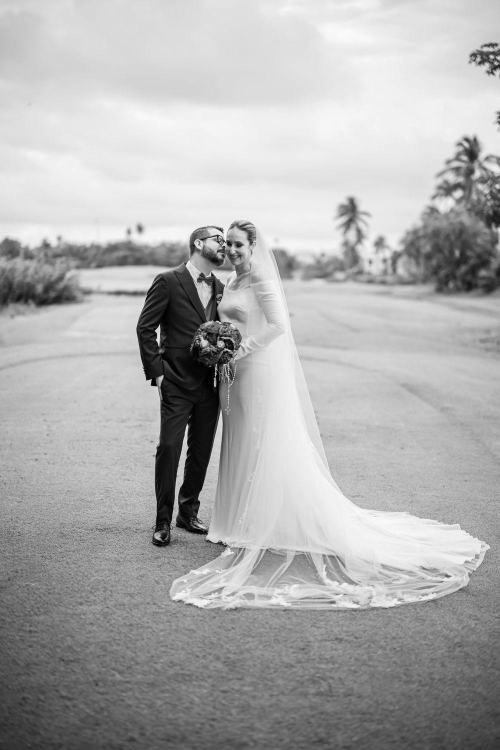 Classic black and white wedding portrait on a palm-lined roadway featuring an elegant long-train bridal gown.