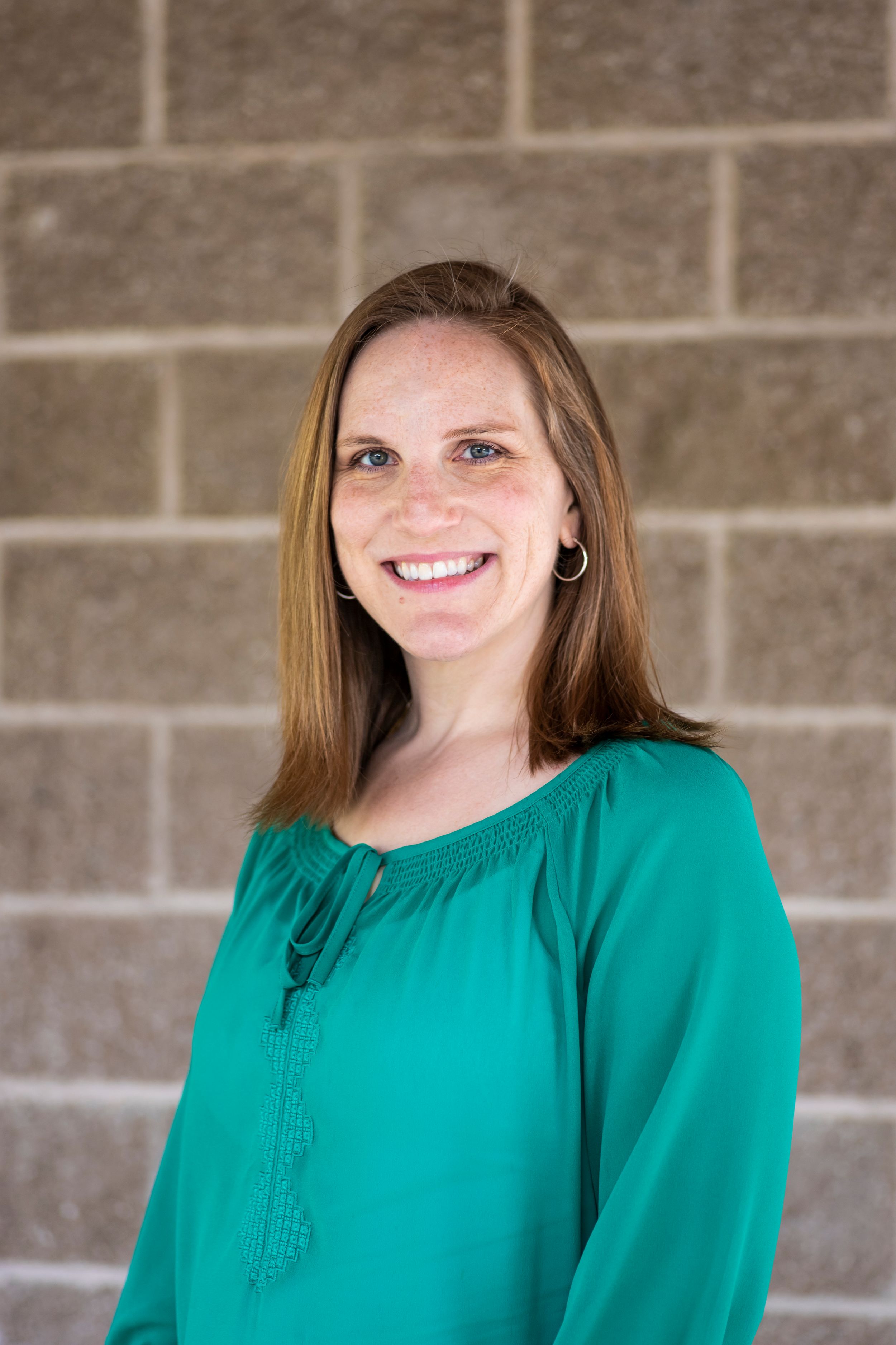 Professional headshot of someone in a green blouse smiling warmly against a stone wall background.