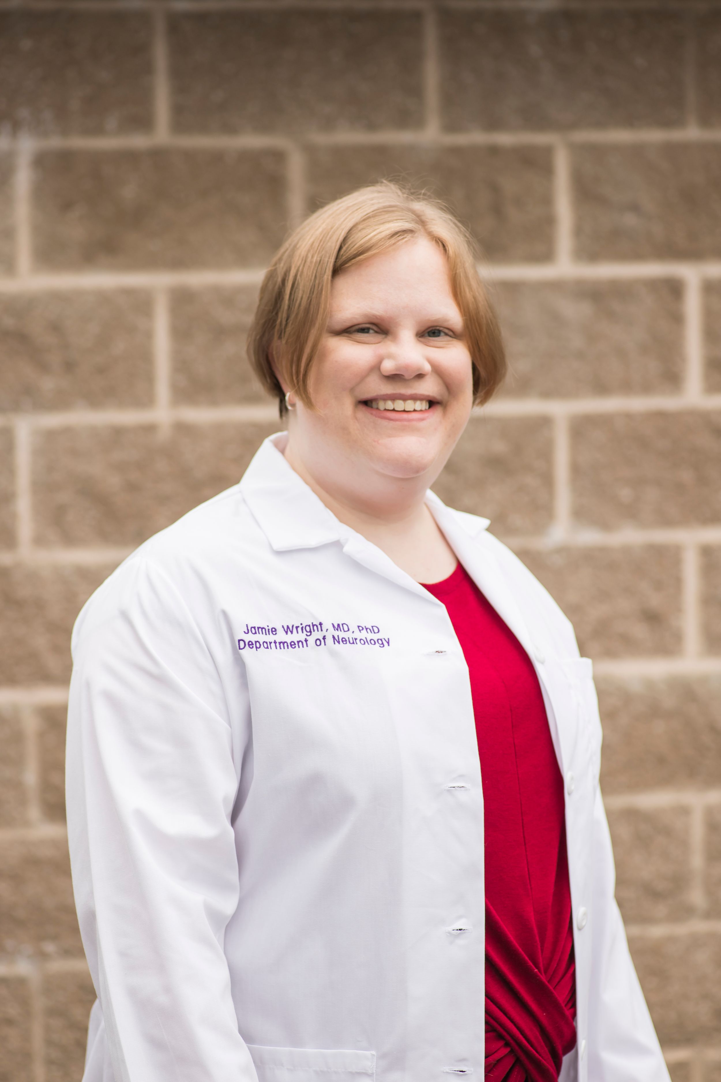 Healthcare professional in white lab coat and red top poses against stone wall with friendly smile.