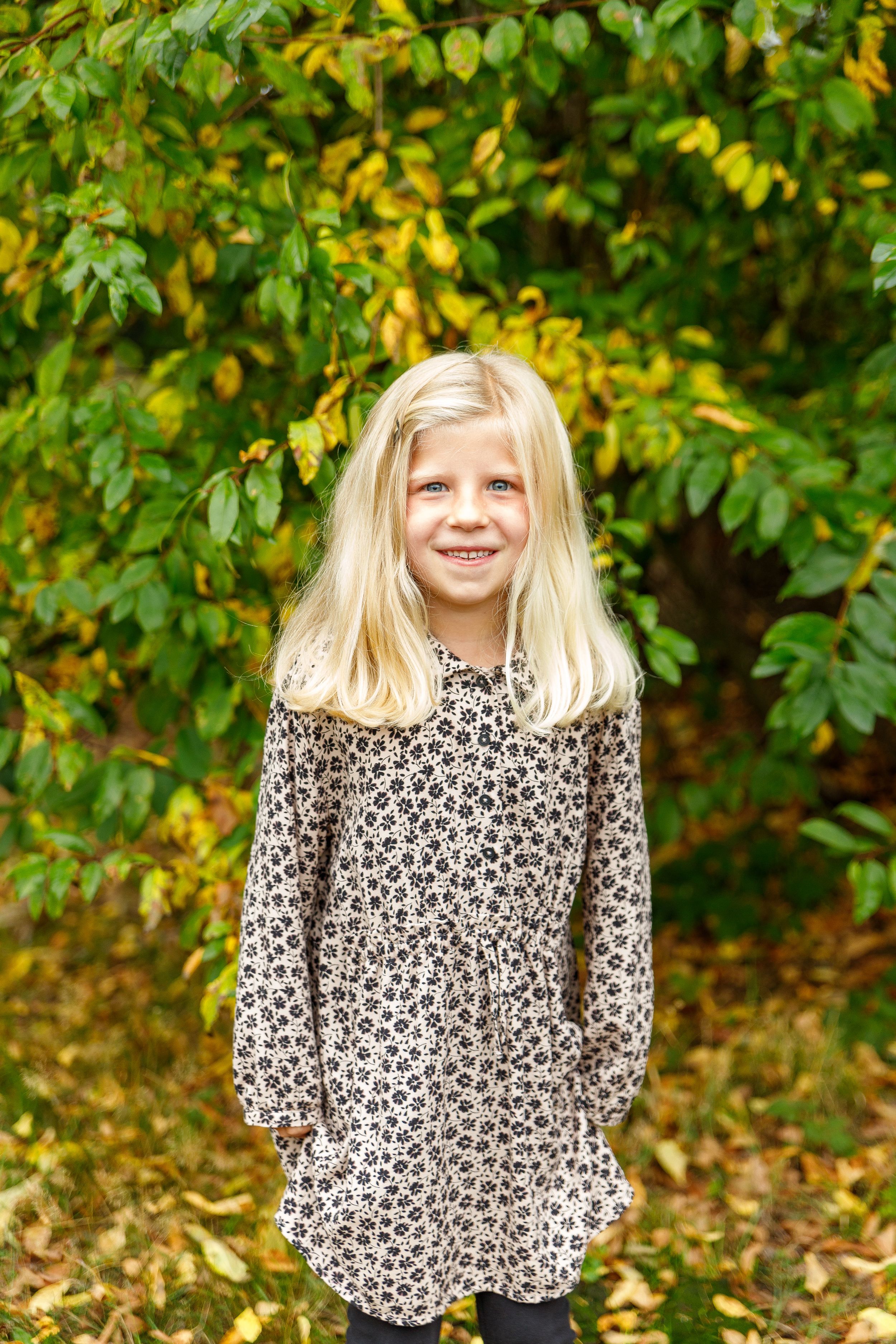Child in floral print dress standing in front of autumn foliage.