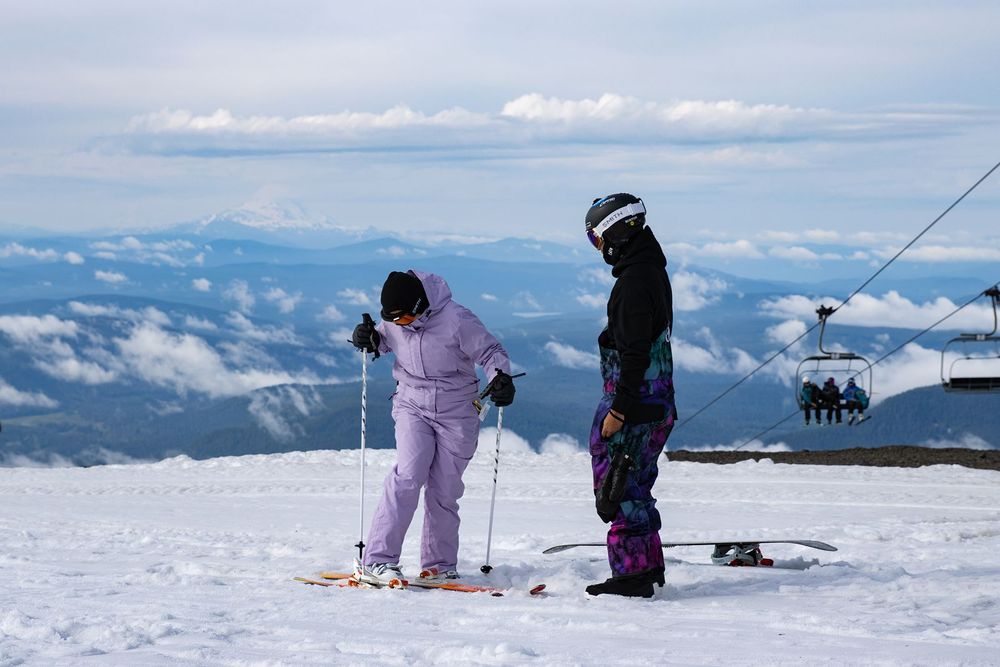 Two skiers in colorful snow gear stand on a snowy mountain summit with ski lift and clouds visible below.