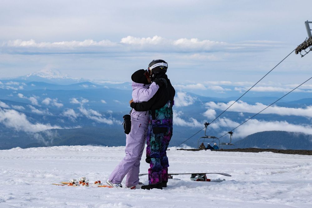 Two snowboarders embrace on a snowy Mt.Hood at Timberline Lodge with clouds and scenic valley views below.