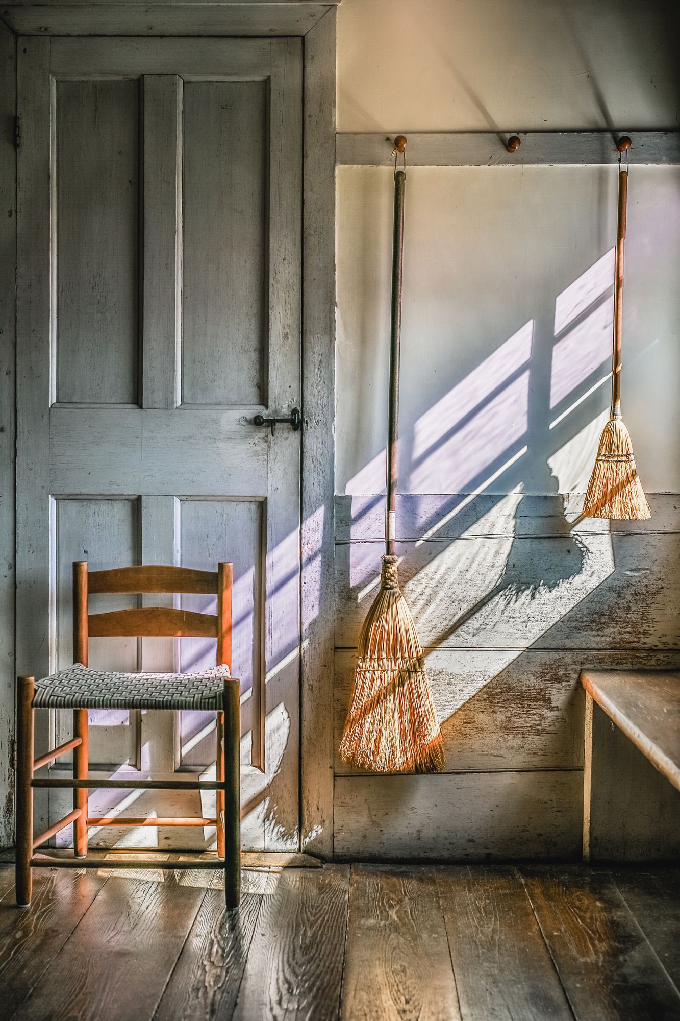 Shaker brooms hang in sunlight streaming through window onto worn hardwood floors at Canterbury Shaker Village in Canterbury, New Hampshire