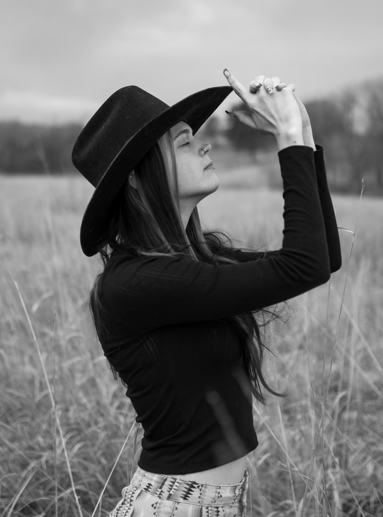 Person in black attire and hat poses dramatically in a misty field.