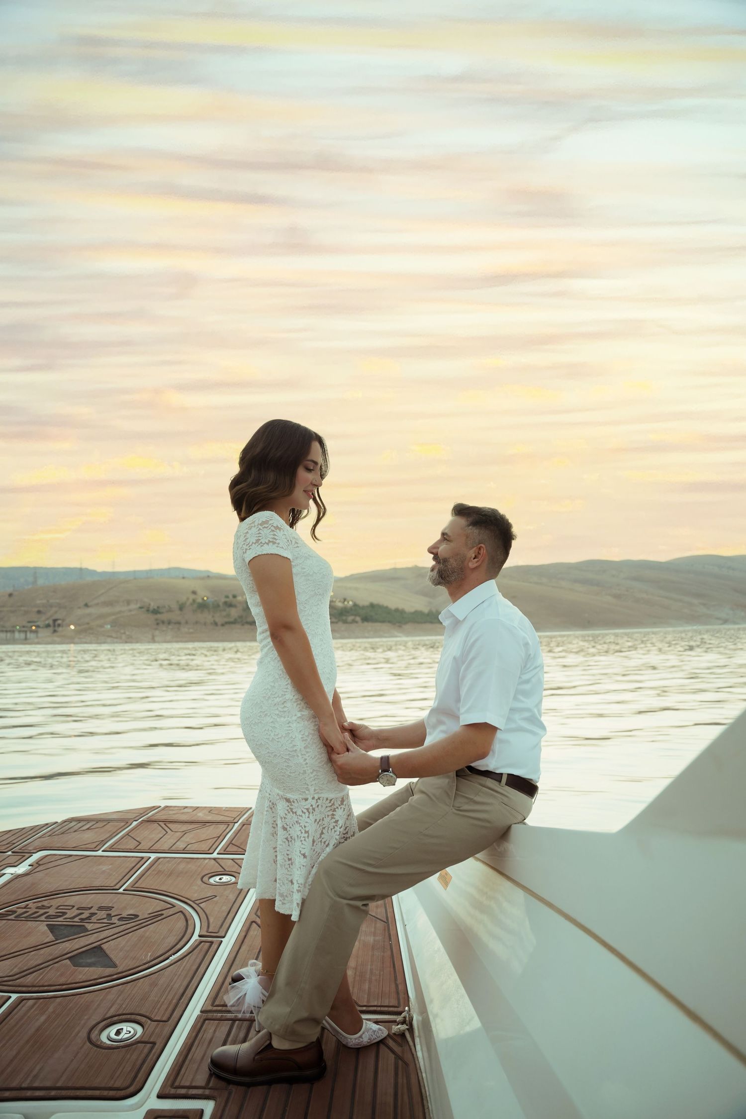 A couple shares an intimate moment on a boat deck at sunset with mountains and water in the background.