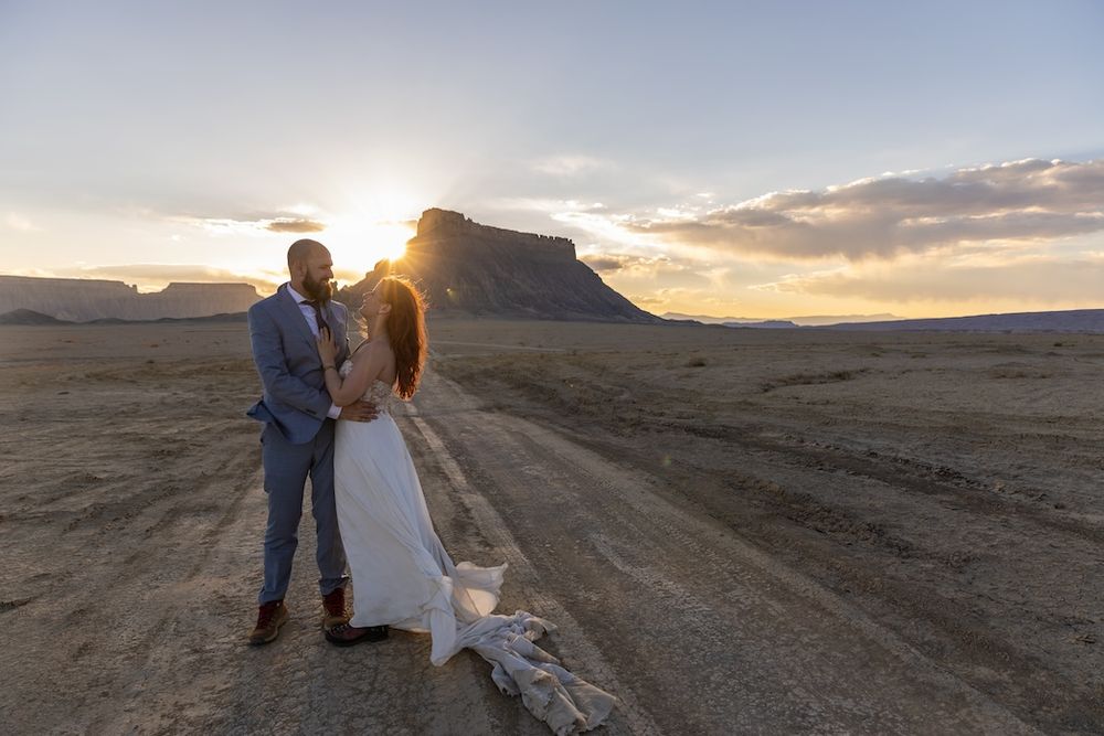 A couple in formal attire poses together on a dirt road at sunset in a desert landscape.