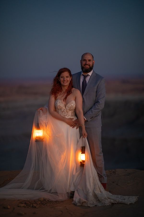 Romantic beach wedding photos at dusk with glowing lanterns casting warm light on a couple in formal attire.