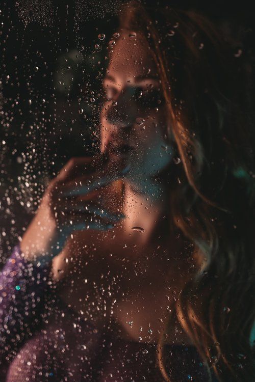 Artistic portrait shot through wet shower with colorful lighting effects creating moody atmospheric ambiance.