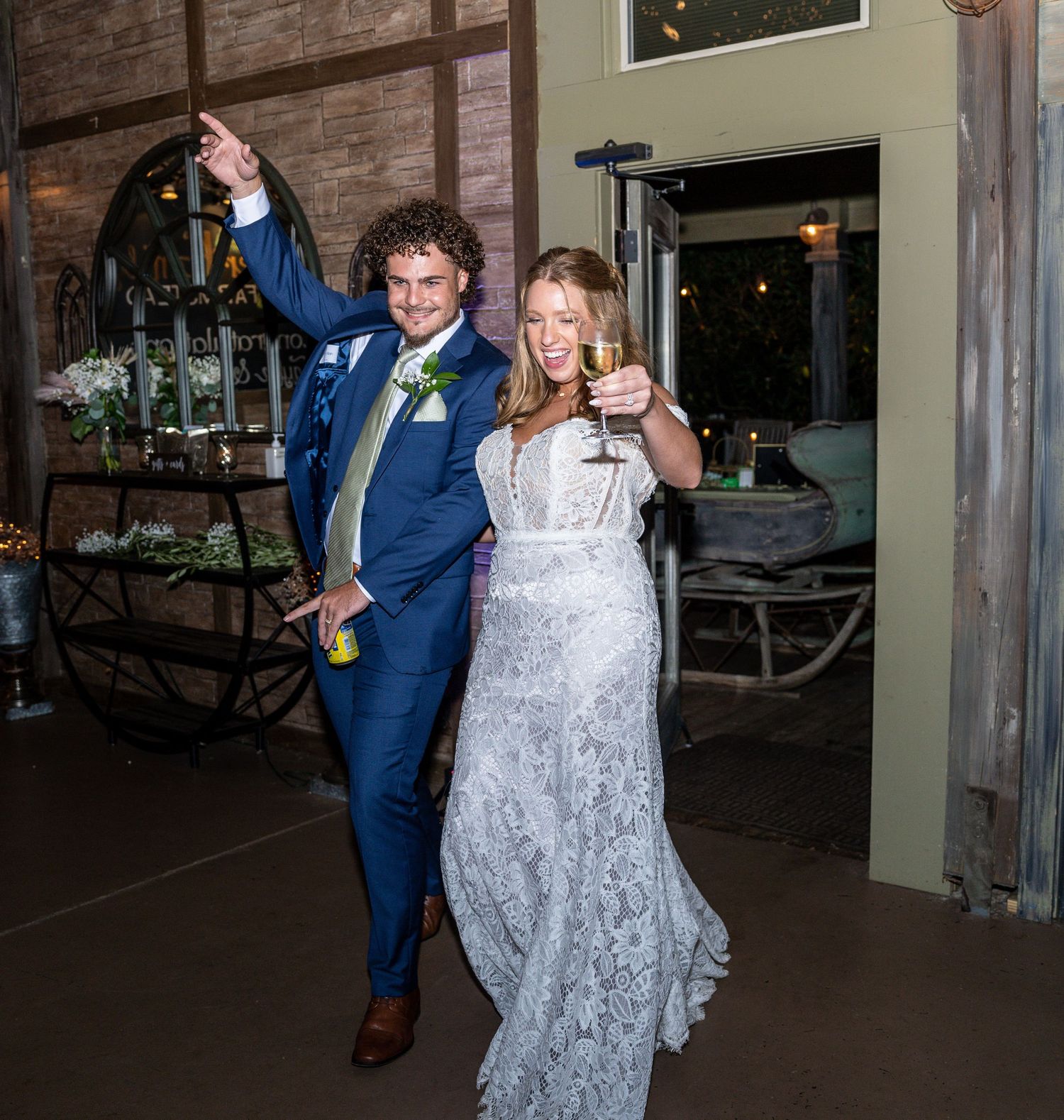 Celebratory pose of newlyweds in blue suit and lace dress at rustic venue.