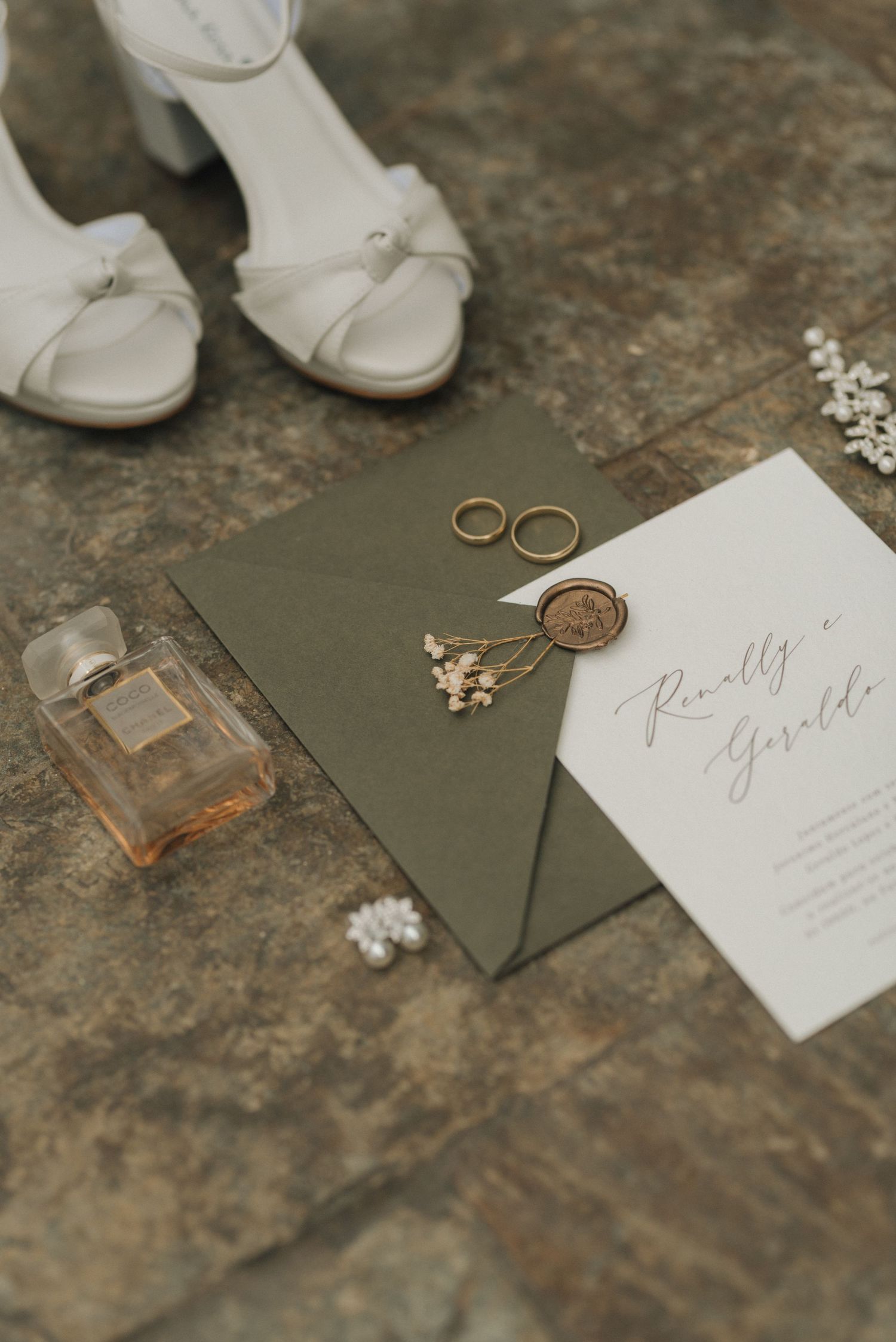 Wedding details with white shoes, rings, perfume and invitation on stone surface.