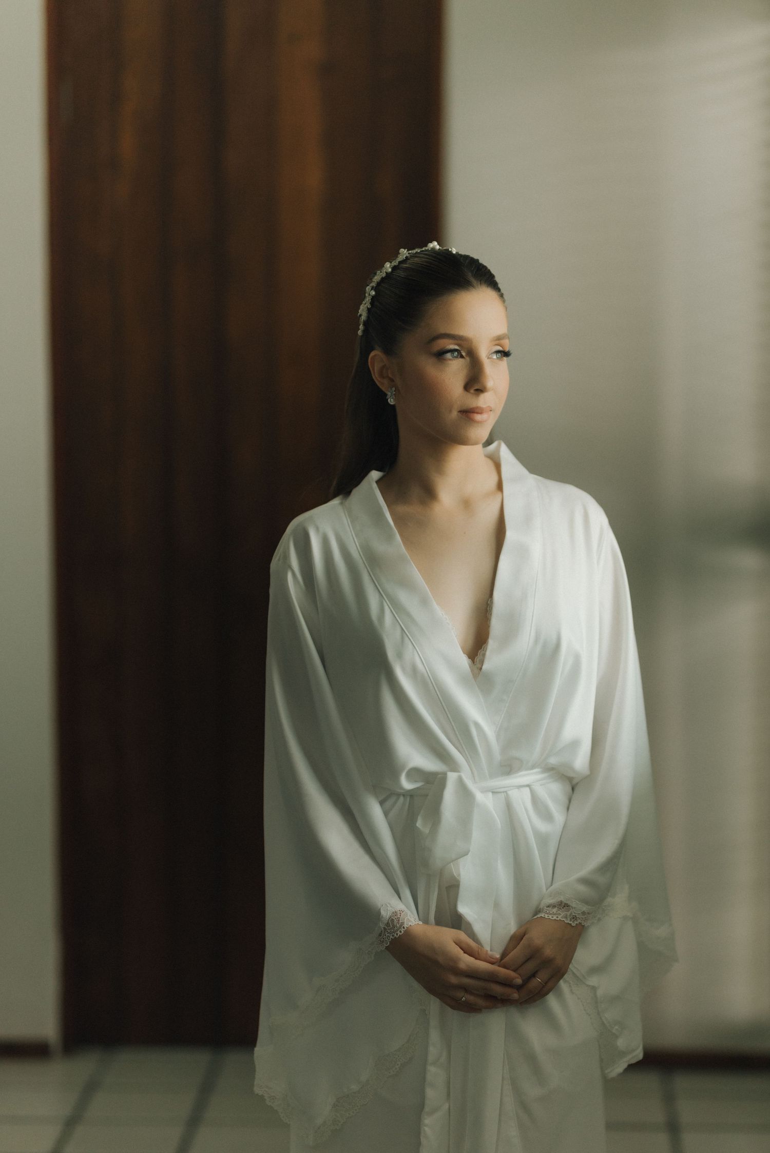A figure in a white silk robe stands elegantly near a window in preparation for a special day.