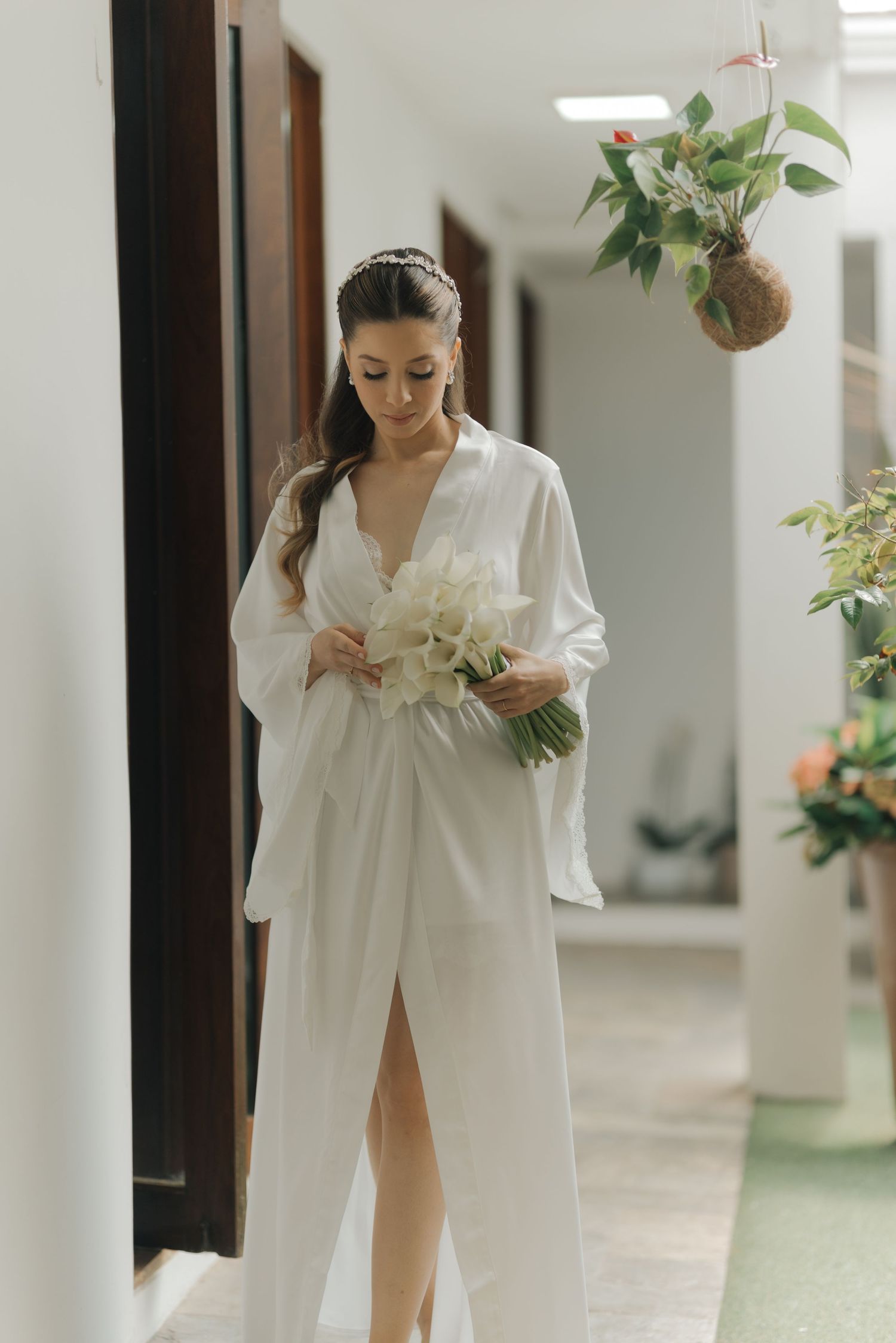 Figure in white robe holds bouquet of flowers in bright interior hallway.