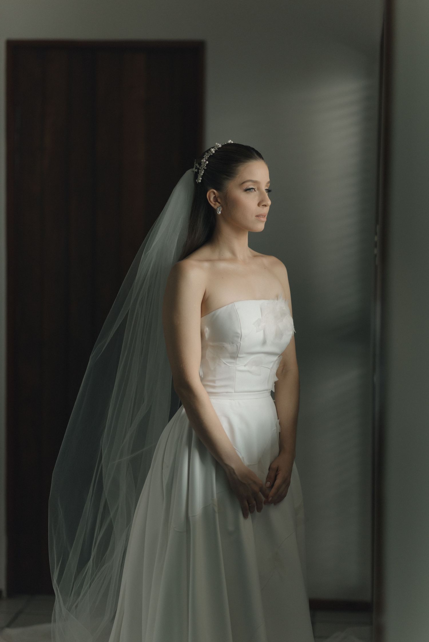 A bride in a classic strapless white gown and veil poses elegantly by a window in soft natural light.