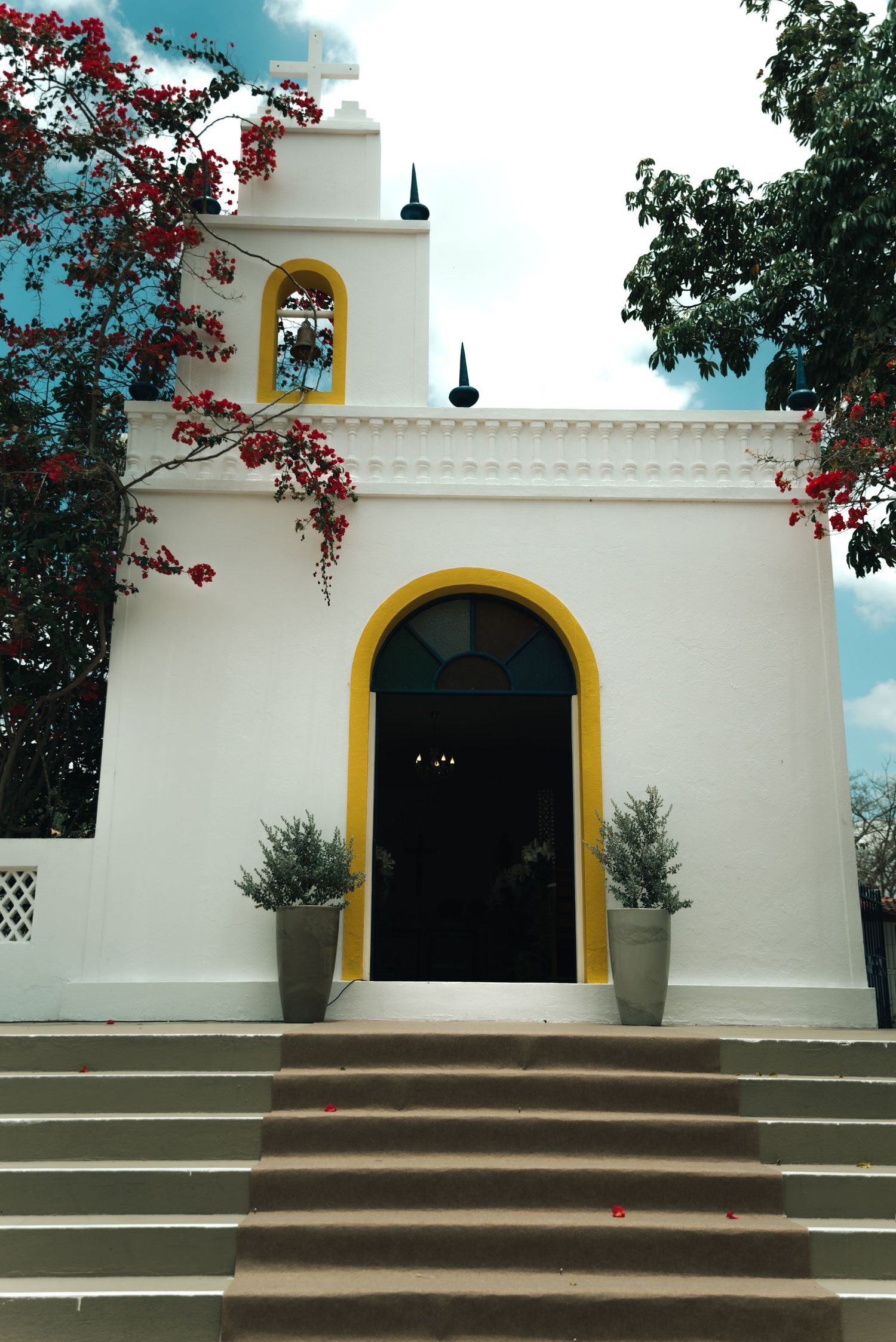 A white church with yellow trim and arched doorway features flowering bougainvillea and stone steps.