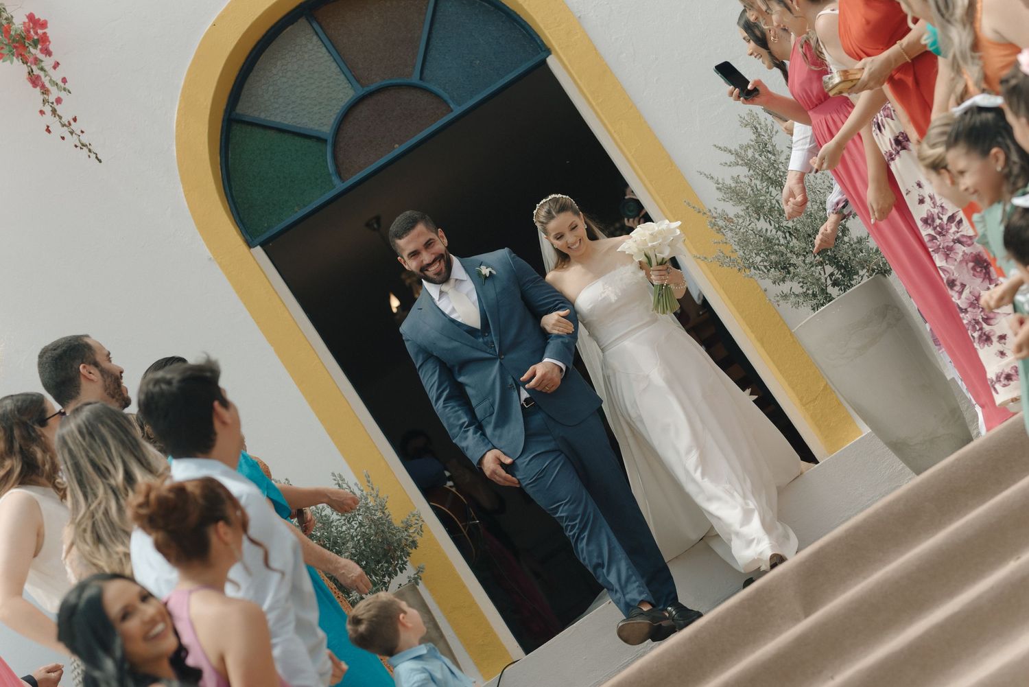Newlyweds exit a church through an arched doorway as guests look on from the steps.