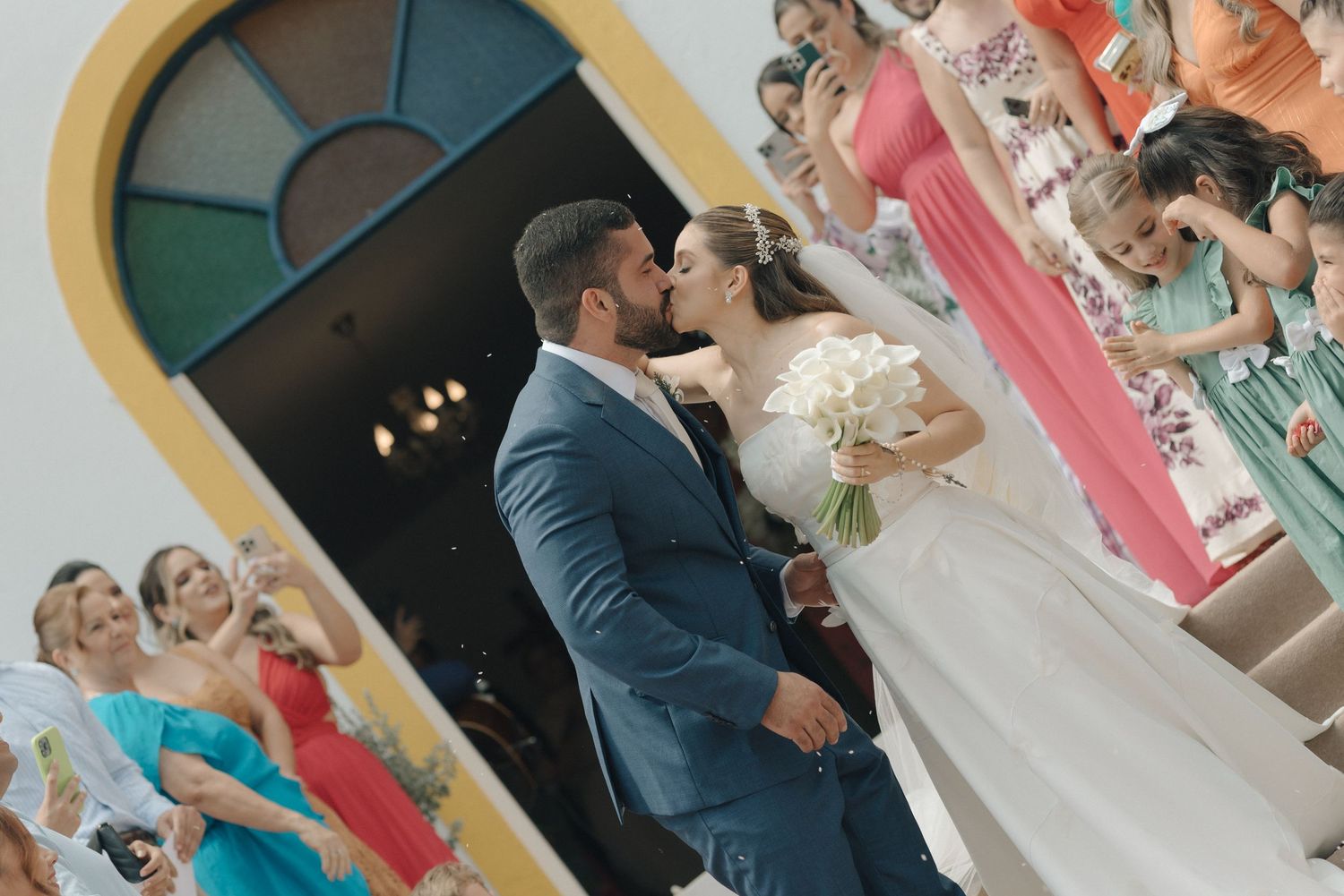 A romantic kiss between newlyweds at their church exit while guests look on.