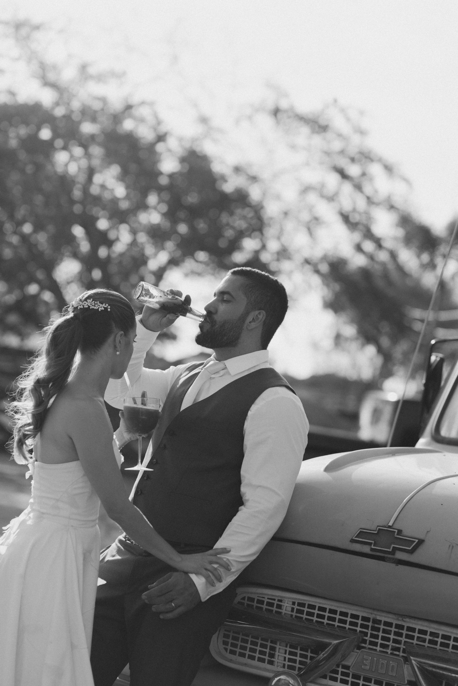 Black and white portrait of newlyweds sharing drinks by vintage Chevrolet truck.