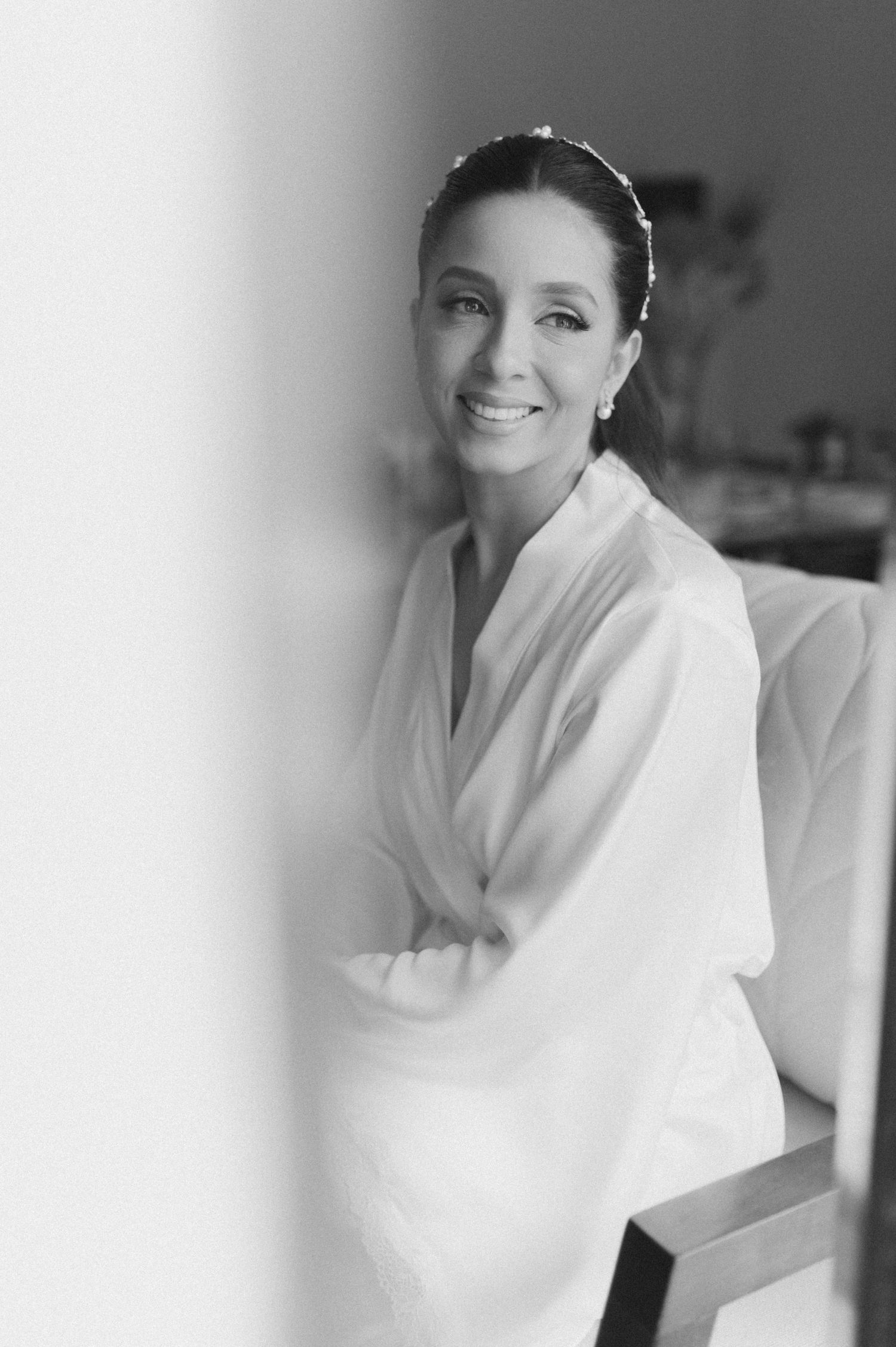 A bride in preparation wearing a white robe smiles in a black and white portrait.