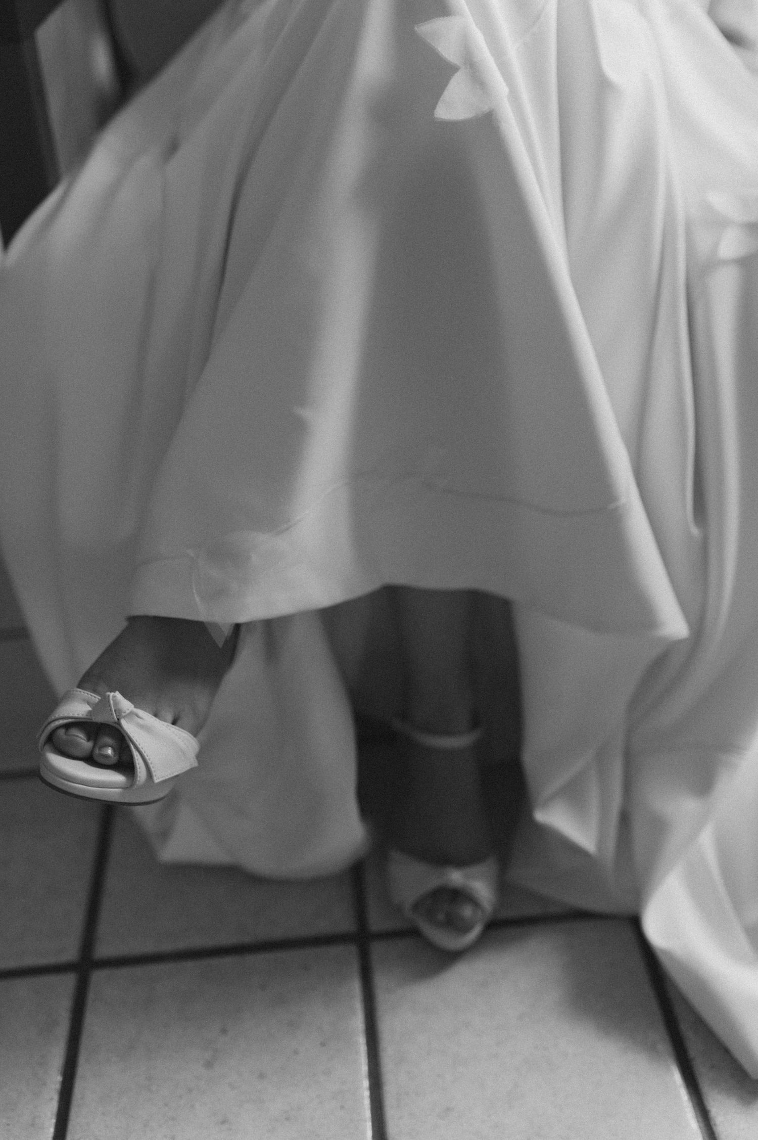 Black and white detail shot of bride's feet in elegant shoes beneath wedding dress hem.