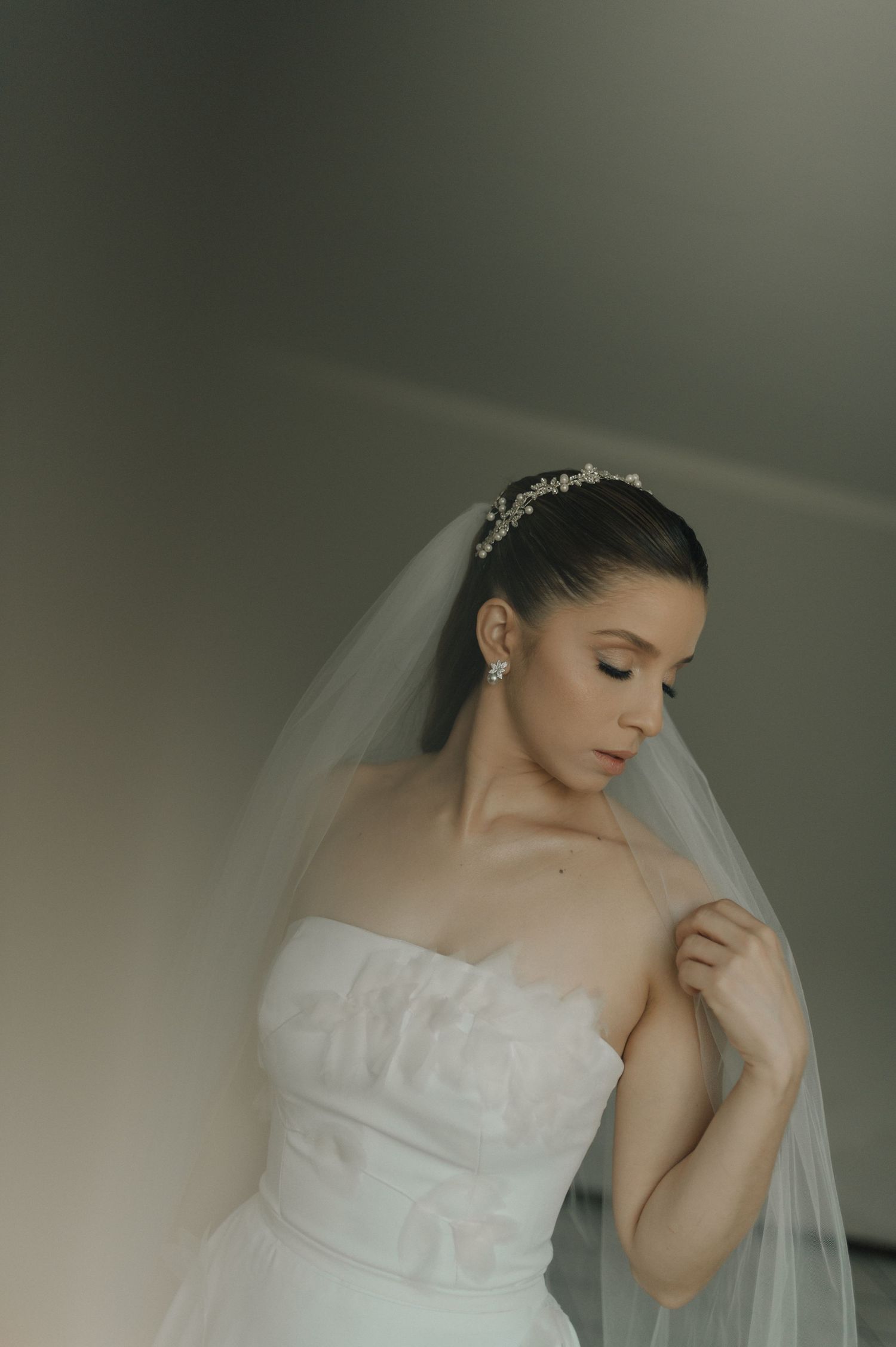 A bride in a white strapless wedding gown and veil poses elegantly with delicate jewelry and soft lighting.