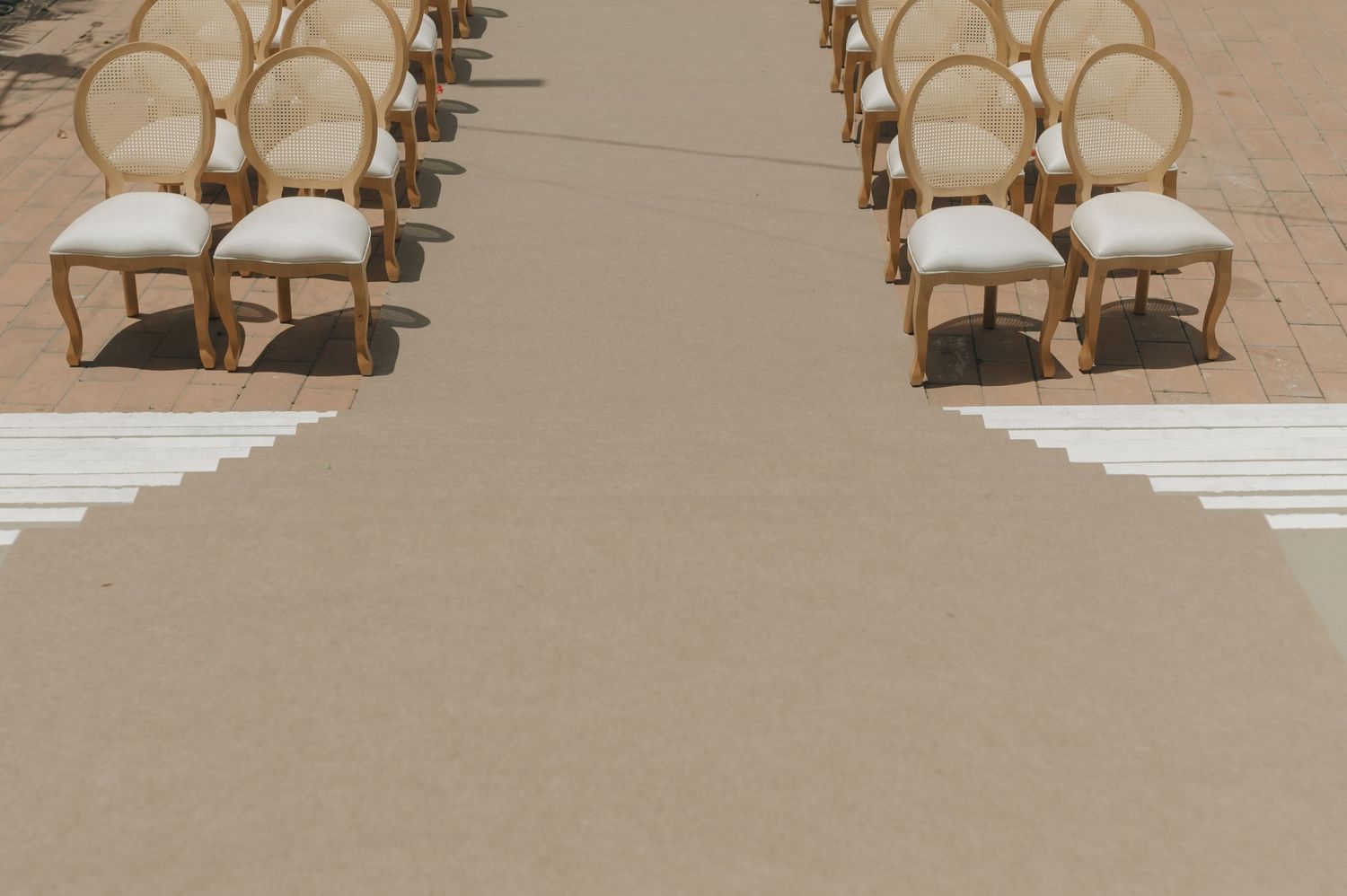 Rows of elegant cream-colored chairs line an aisle at an outdoor wedding venue.