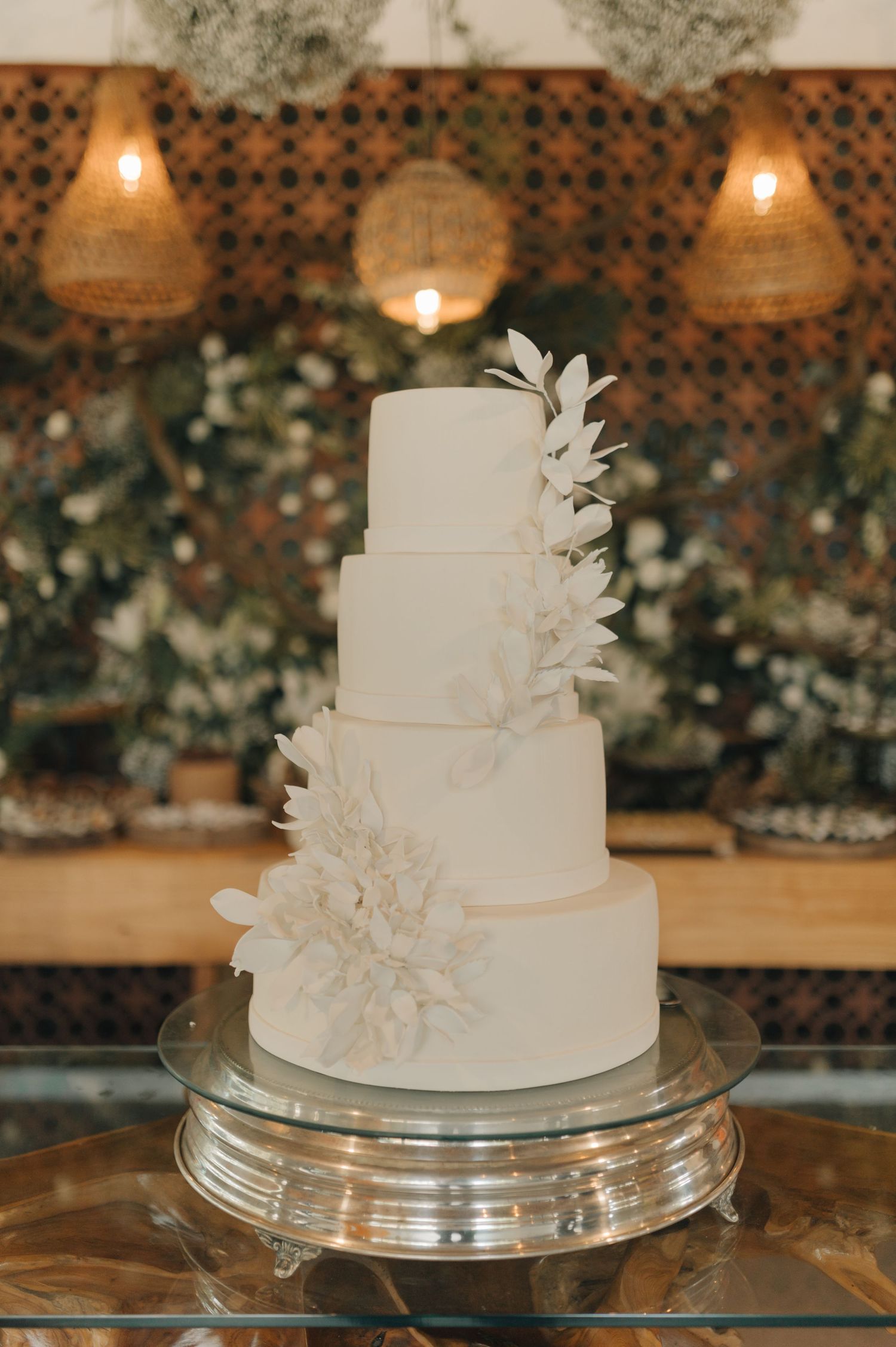 Four-tiered white wedding cake decorated with delicate floral details and illuminated by hanging lights.