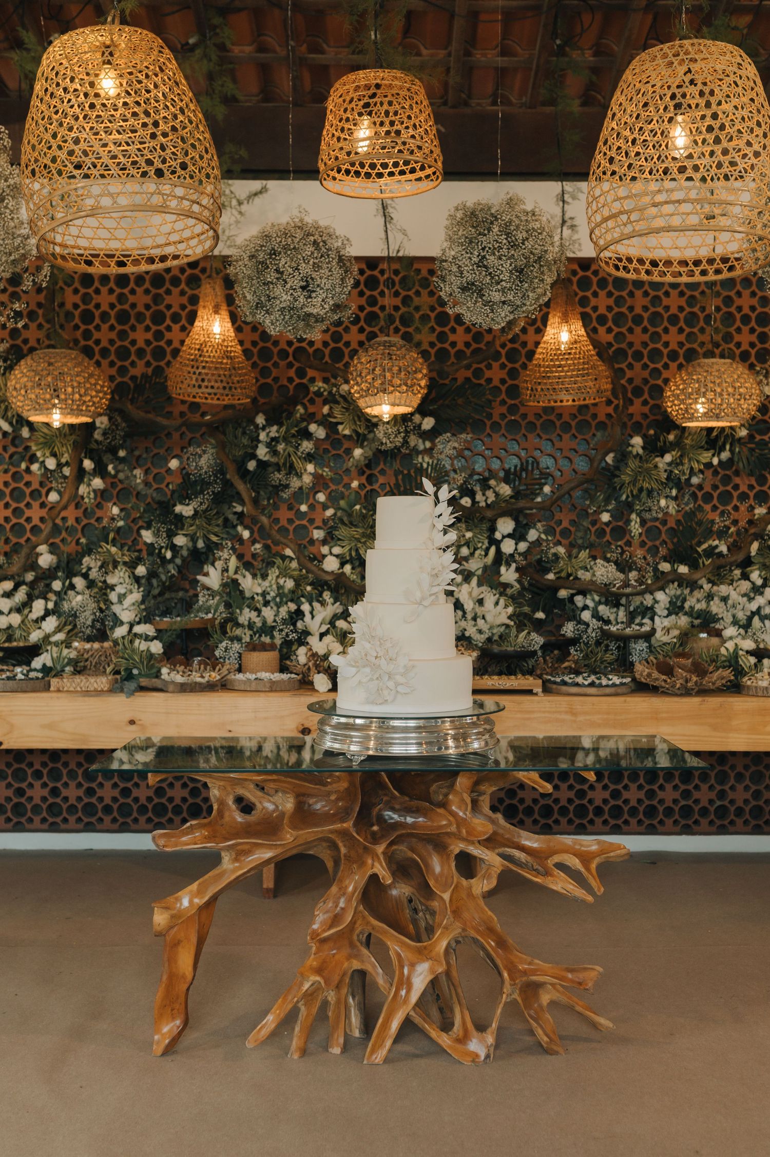 Wedding cake display with woven bamboo pendant lights and floral backdrop on a unique driftwood table.