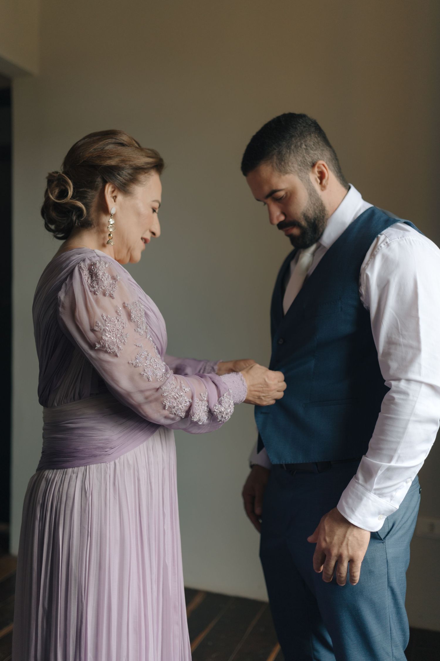 Two people share an intimate getting-ready moment, one adjusting the other's formal attire.