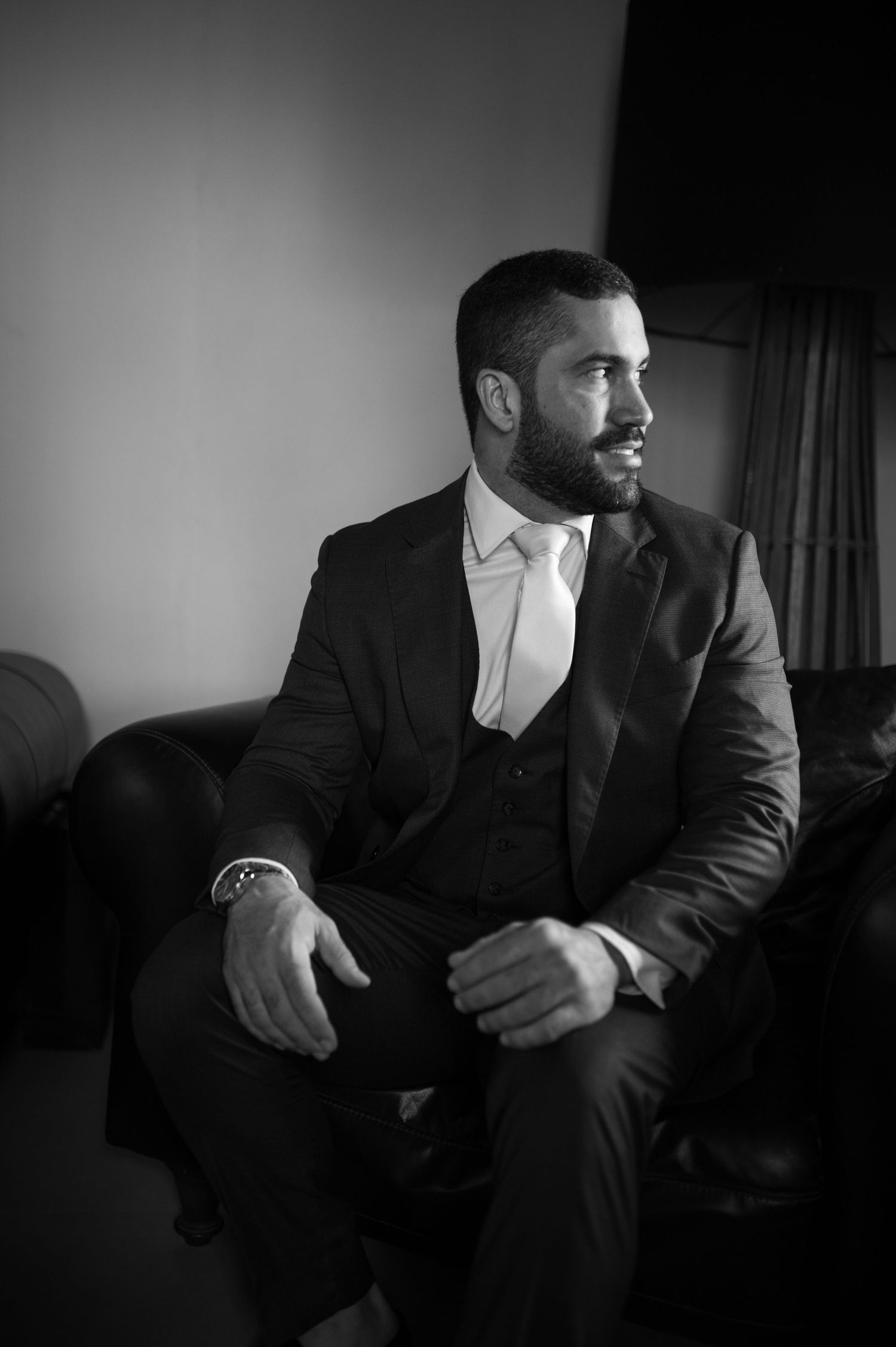 A formal portrait of someone in a dark suit sitting on a couch looking to the side.